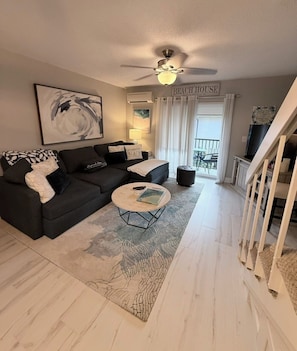 Un salon avec un canapé foncé, une table basse ronde, un ventilateur de plafond et une enseigne indiquant « MAISON DE PLAGE ».