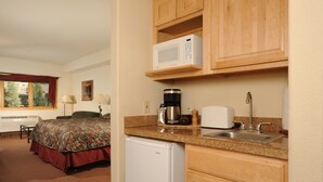 A compact hotel room with a kitchenette, a bed with a floral bedspread, a window with a view of trees, and a small refrigerator. - Cozy Studio Suite | Pool, Hot Tub & Parking! (Keystone)