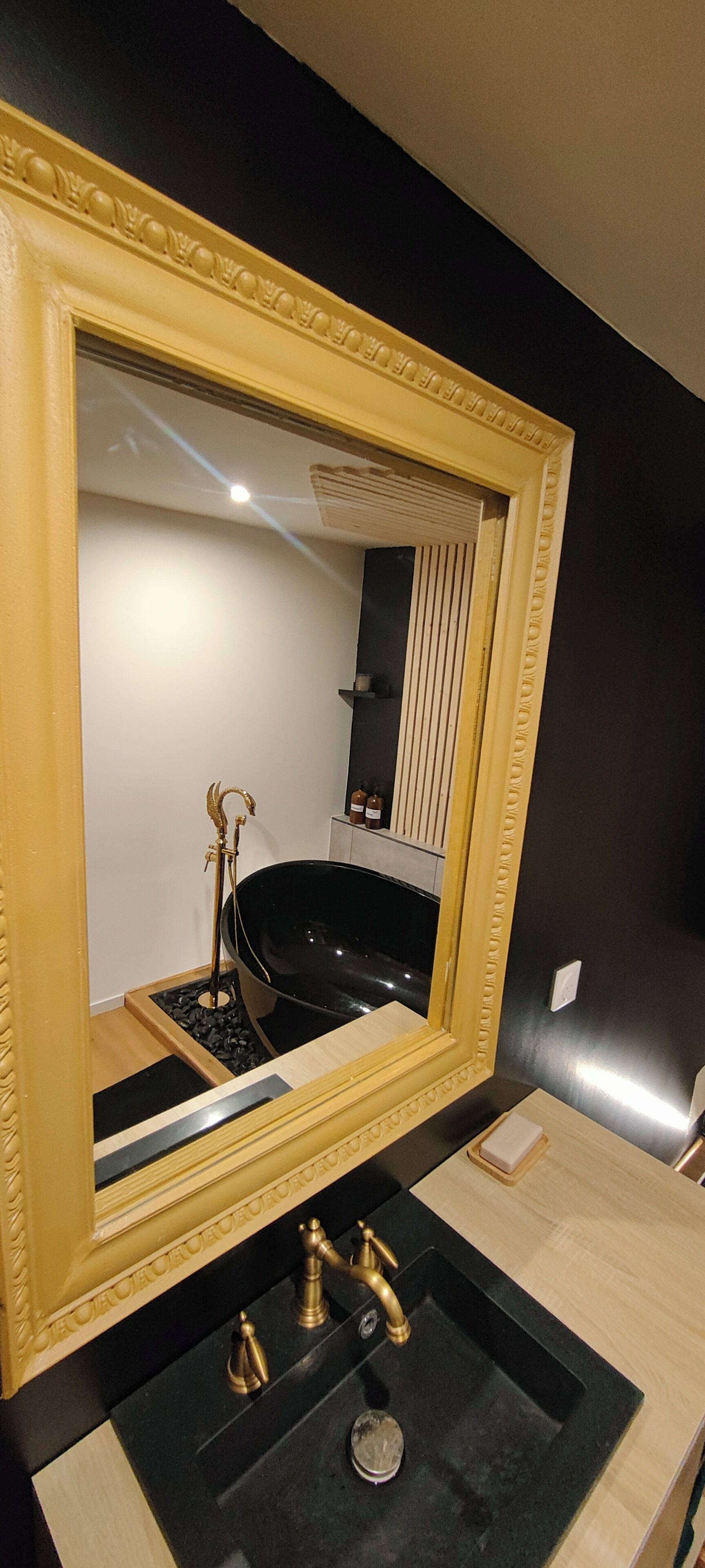 A bathroom with a large mirror, black sink, gold faucets, and a bathtub.