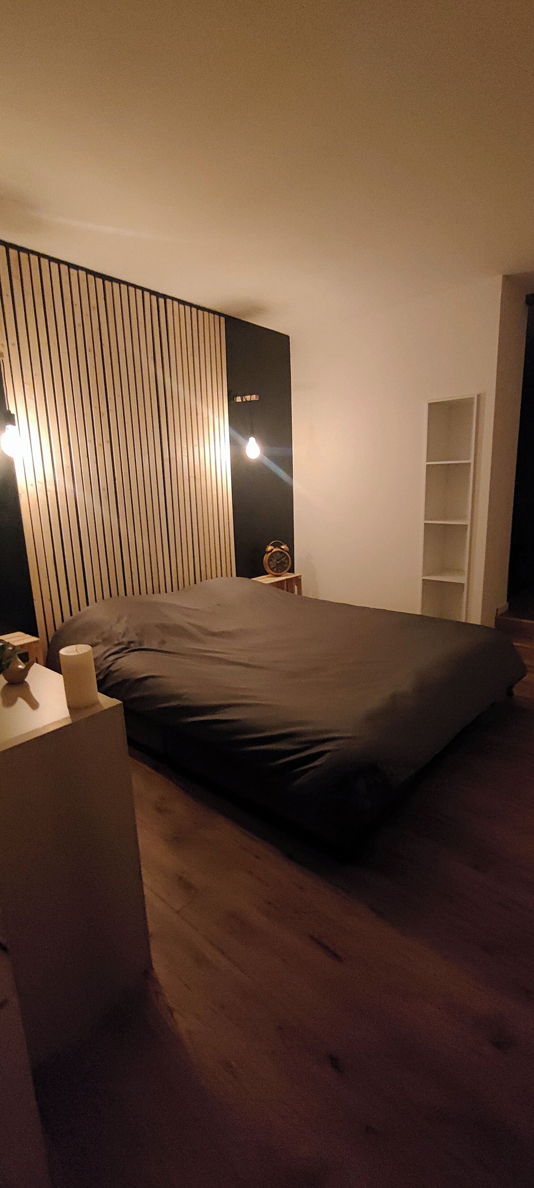 A bedroom with a bed, bedside table, lamp, and a wooden slat wall.