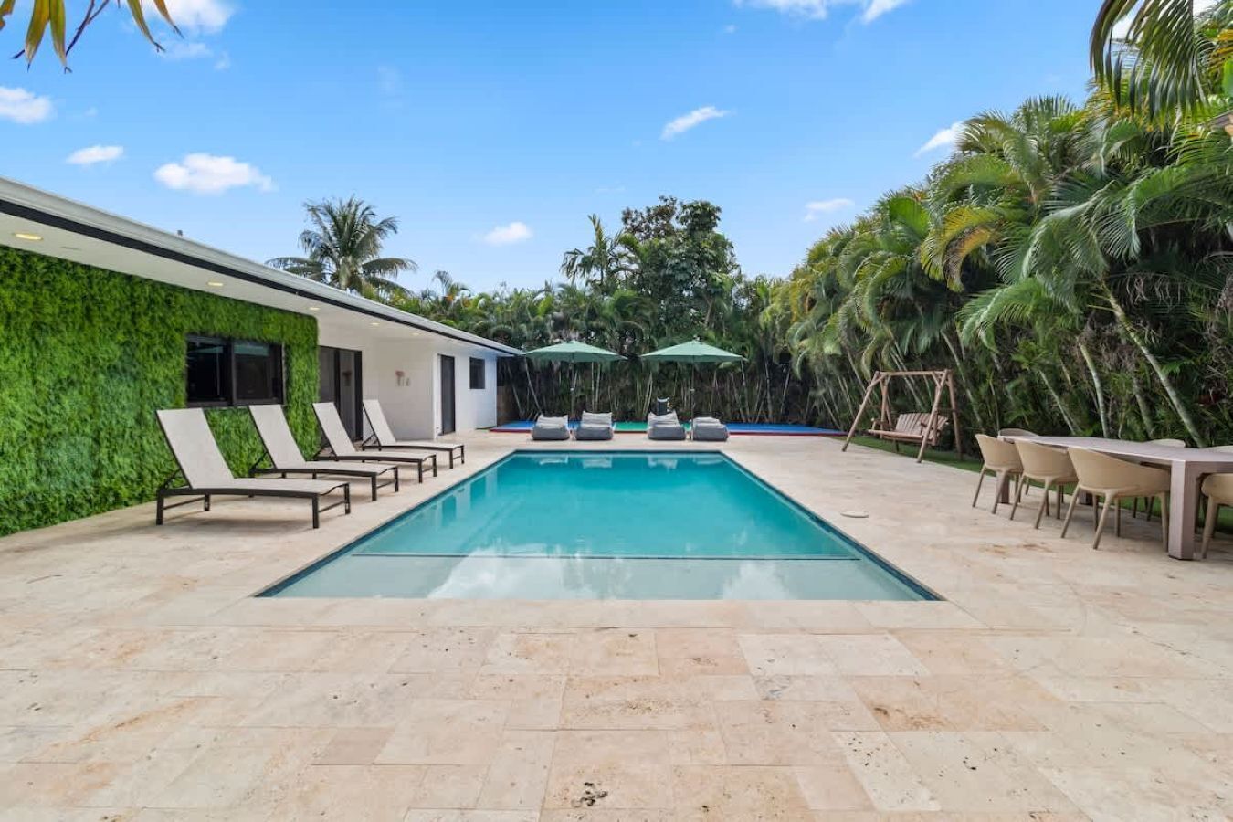 A swimming pool with lounge chairs and a table, surrounded by palm trees and a green wall.