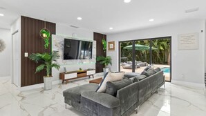 Ein modernes Wohnzimmer mit einem grauen Ecksofa, einem Flachbildfernseher, einer Wandpaneele aus Holz und einem großen Fenster mit Blick auf einen Poolbereich.