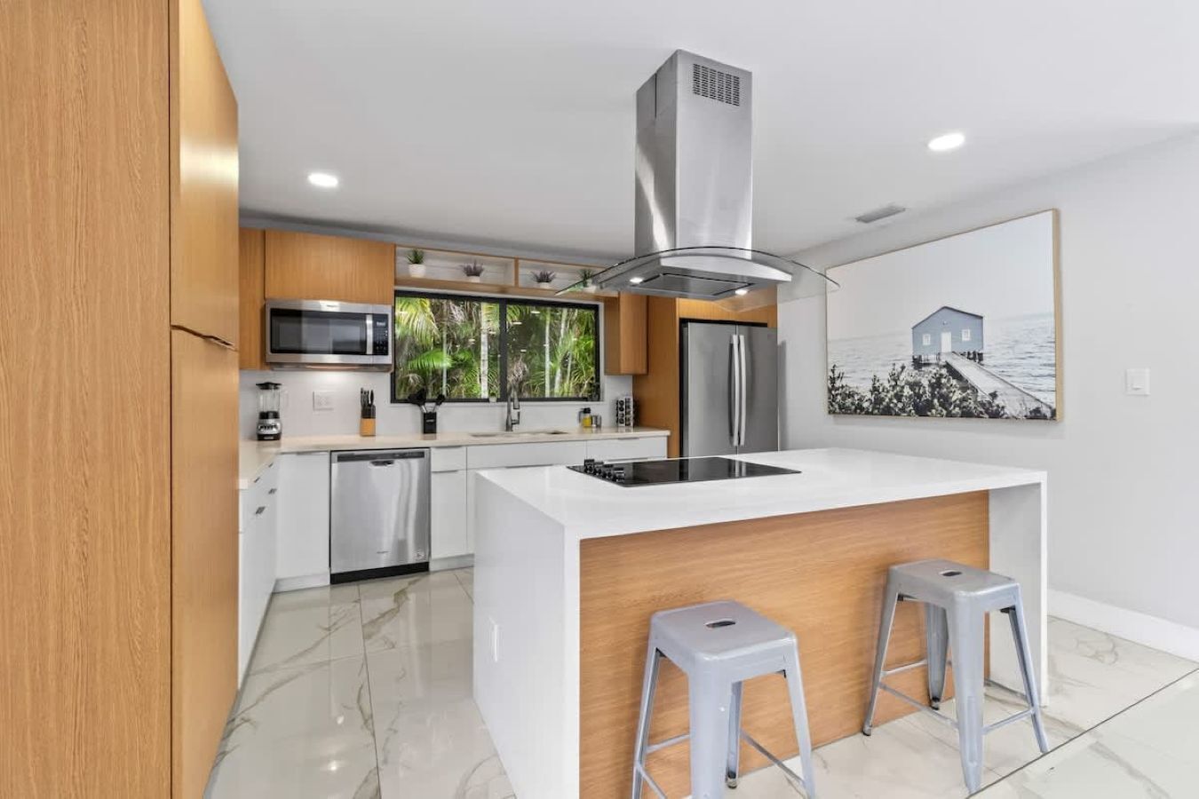 Une cuisine moderne dotée d’un îlot central, d’appareils électroménagers en acier inoxydable, de meubles en bois et d’une grande fenêtre offrant une vue sur la verdure.