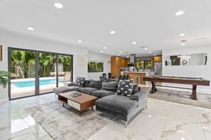 A modern living room with a pool, grey sofa, coffee table, and a pool table. - Heated Pool Villa in Miami by Glam Homes (North Miami Beach)