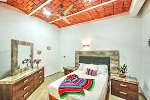 Ein Schlafzimmer mit einem hölzernen Kopfteil, einem Bett mit weißer Bettwäsche, einer farbenfrohen Decke, einem Nachttisch mit Pflanze und einem Spiegel auf der Kommode.