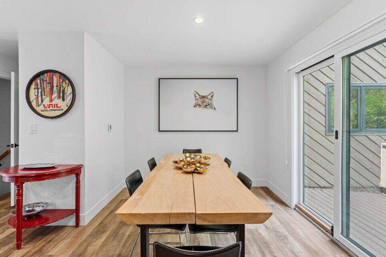 Un comedor con una mesa de madera, sillas, una mesita auxiliar roja, un cuadro enmarco y una puerta de vidrio que da al exterior.