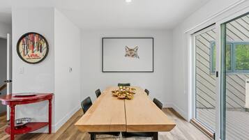 Un comedor con una mesa de madera, sillas, una mesita auxiliar roja, un cuadro enmarco y una puerta de vidrio que da al exterior.