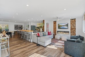 A modern living room with a grey sofa, a dining area with bar stools, a fireplace, and a TV mounted on the wall. - The Alpine Atelier I 4BR Townhome Near Shuttle Stop (Vail)