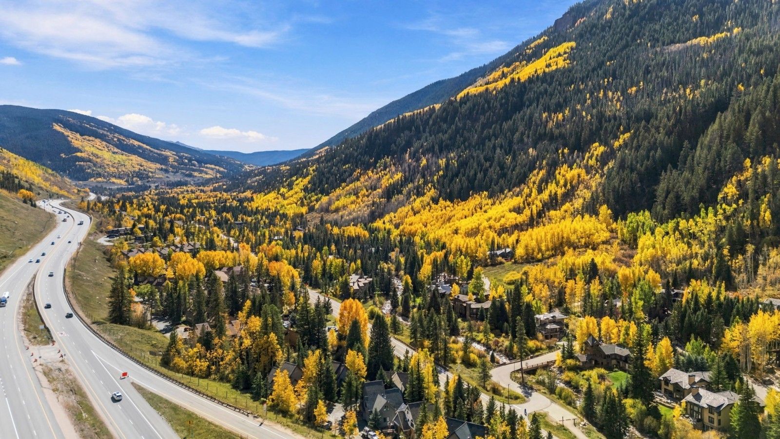 Una carretera serpentea por una zona montañosa con árboles de colores otoñales y edificios residenciales.