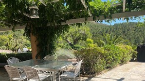 Un patio exterior con mesa y sillas, rodeado de follaje y una pérgola.