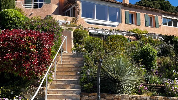 Una casa con escaleras de piedra, un ventanal grande y un jardín con diversas plantas.