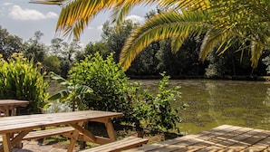 A wooden picnic table with benches, a palm tree, and a body of water in the background. - Cottage Parks La Comtesse (Bélus)