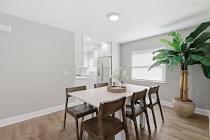 A dining area with a white table, wooden chairs, a potted plant, and a kitchen in the background. - Orange Coast Getaway (Port Orange)