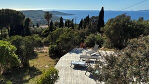 Une terrasse en bois, dotée de chaises longues, qui offre une vue sur un paysage côtier, avec des arbres et la mer à l’horizon.