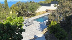 Une terrasse en bois mène à une piscine entourée de chaises longues, le tout étant bordé de verdure et offrant une vue sur une étendue d’eau.