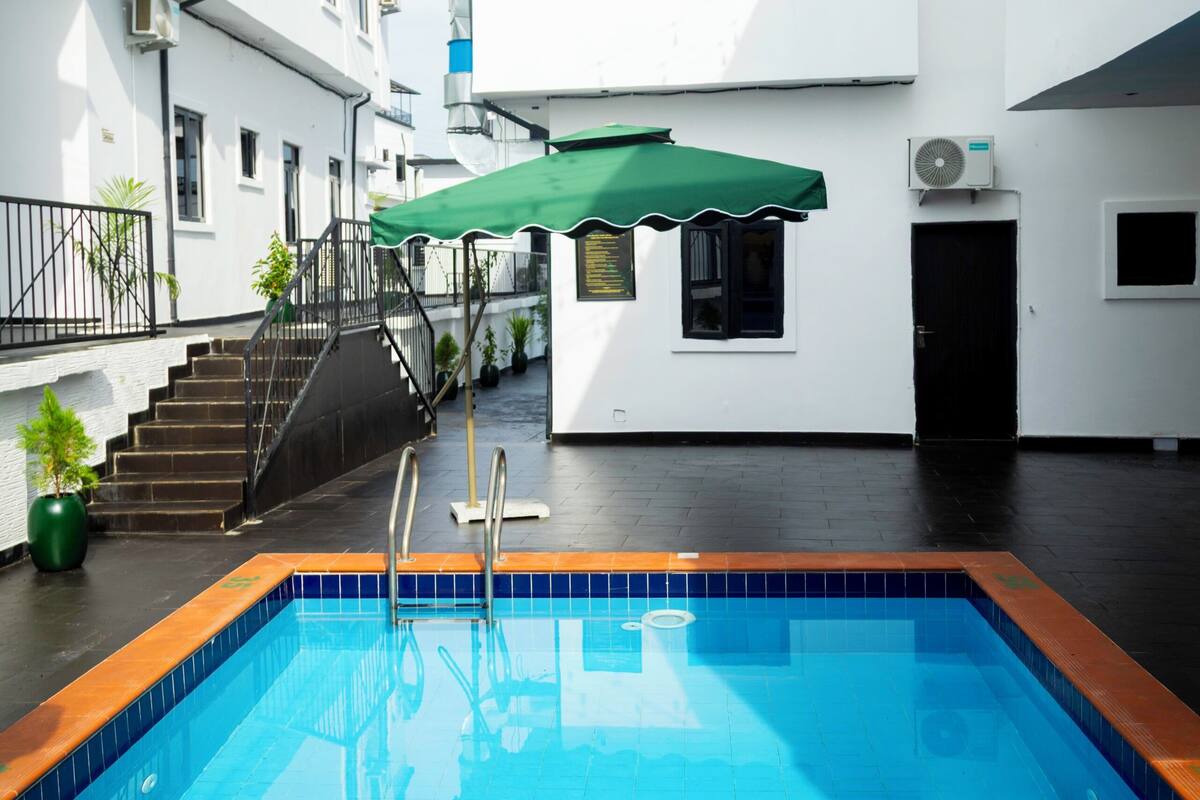 Ein Swimmingpool mit einem grünen Sonnenschirm, einer Treppe und einem Gebäude mit zahlreichen Fenstern und Klimaanlagen.