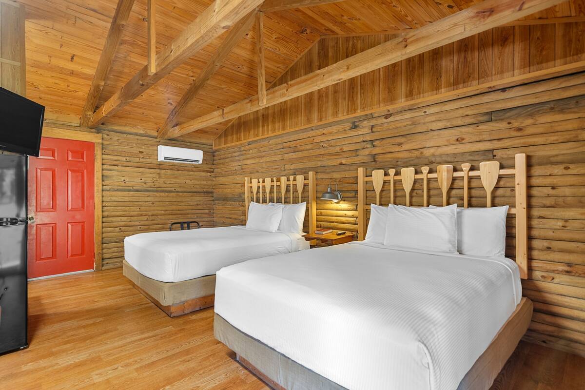 A cabin-style room with two beds, a red door, wooden walls, and a ceiling with exposed beams.