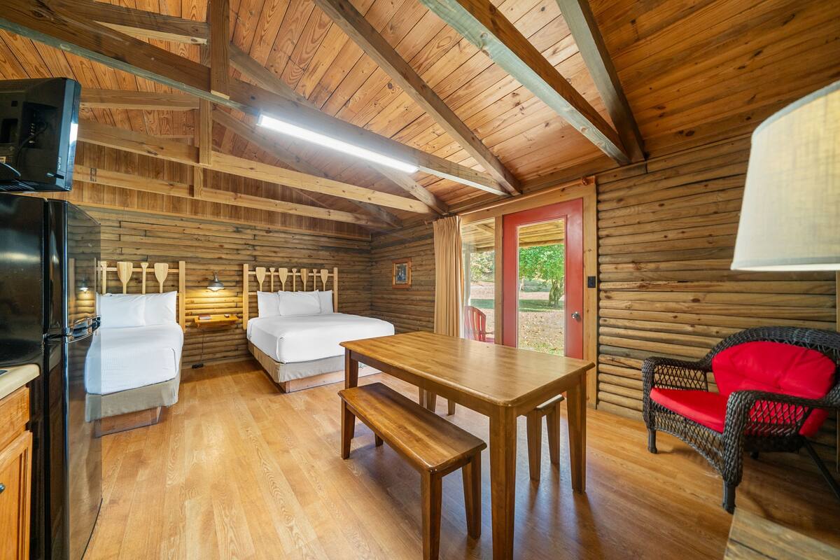 A cabin with wooden walls, a wooden ceiling, a bed, a table, a bench, a chair, and a refrigerator.