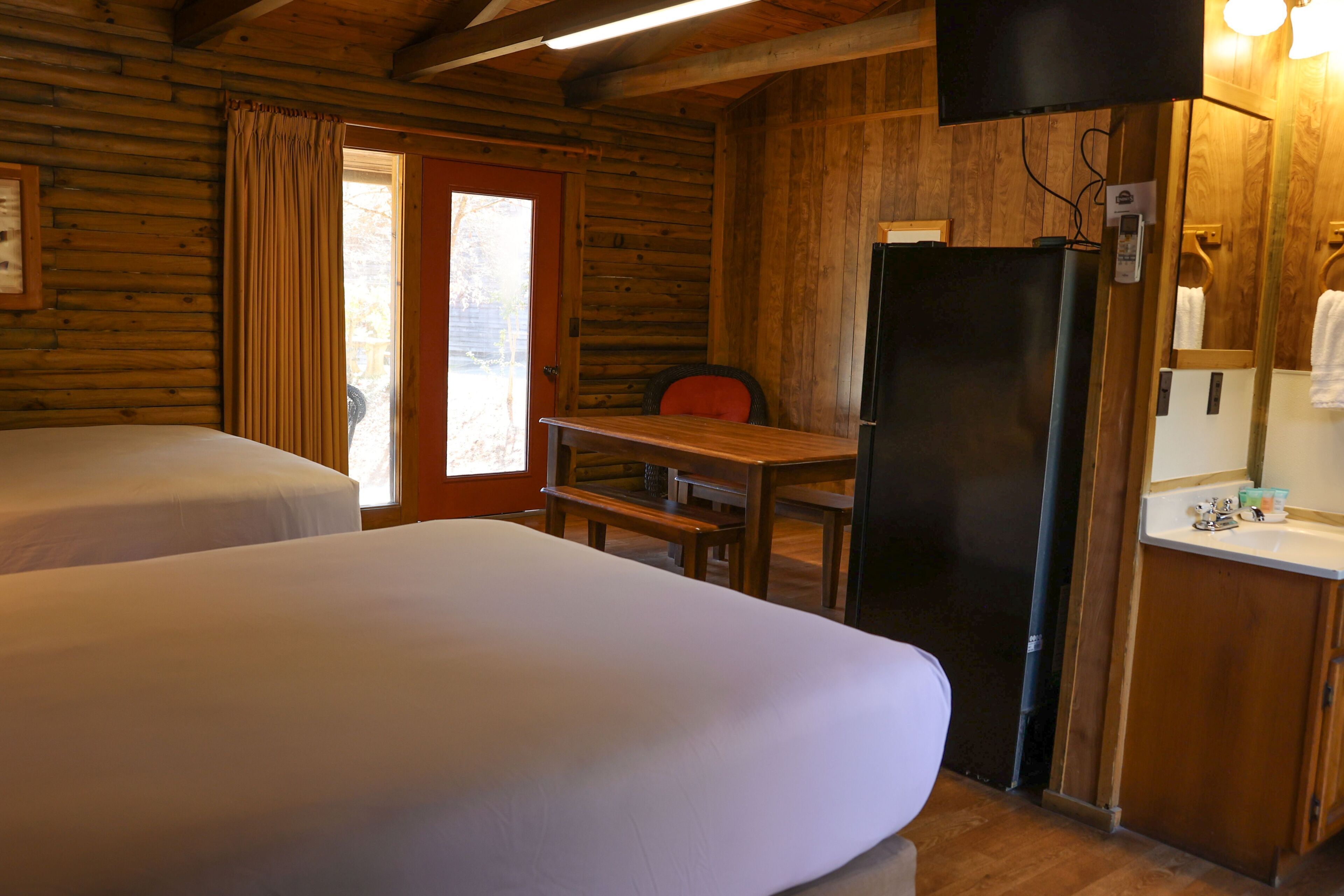 Una habitación tipo cabaña de madera con dos camas, un escritorio de madera, un refrigerador y un baño con espejo.