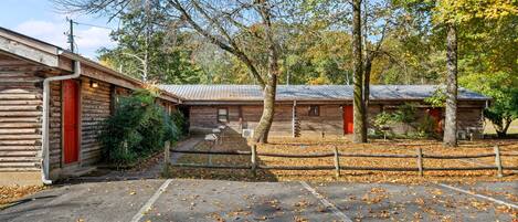 A rustic cabin with a red door, a covered parking area, and a wooden fence.