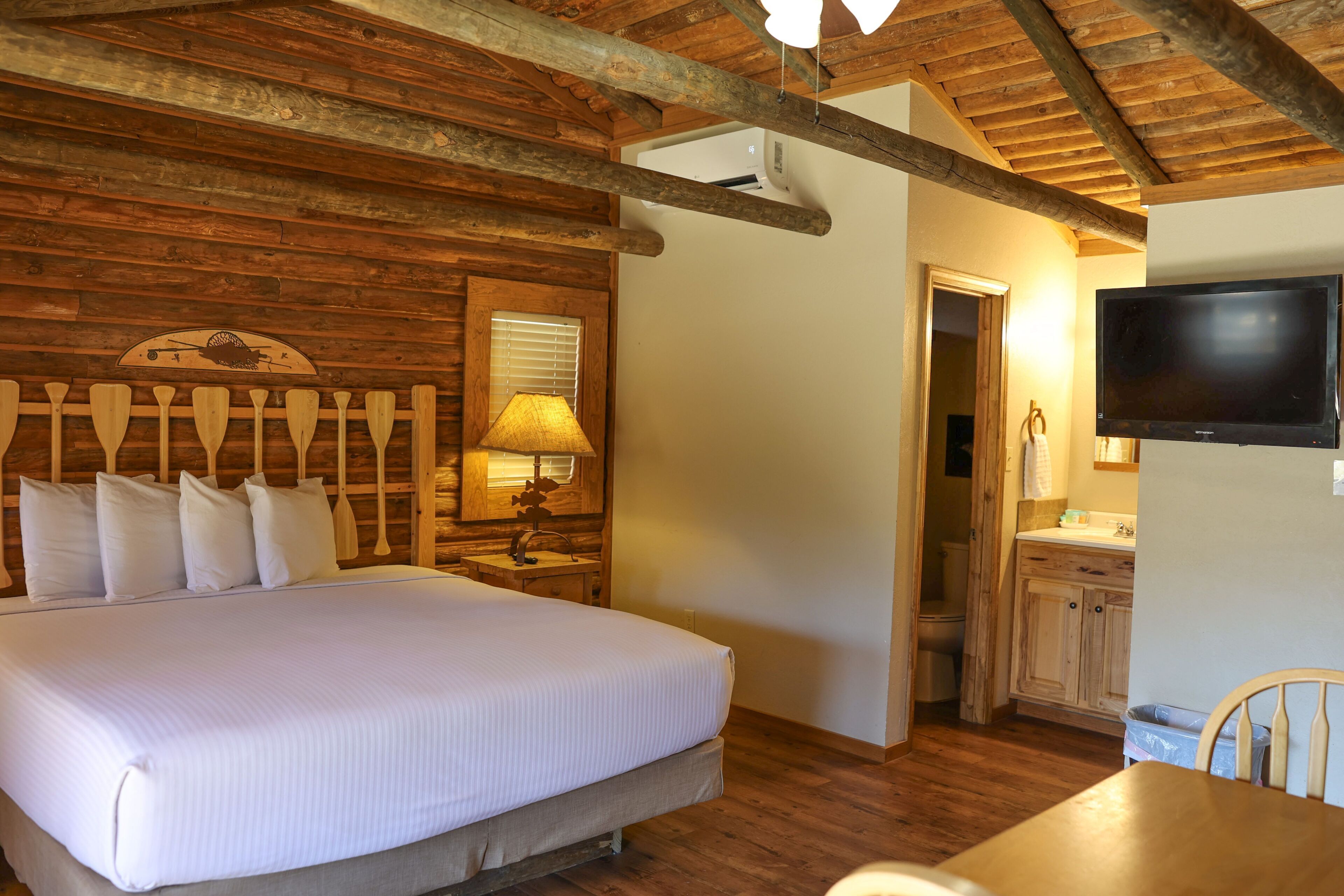 A wooden-paneled bedroom with a bed, a nightstand with a lamp, a television, and a wooden dining table with chairs.