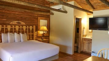 A wooden-paneled bedroom with a bed, a nightstand with a lamp, a television, and a wooden dining table with chairs.