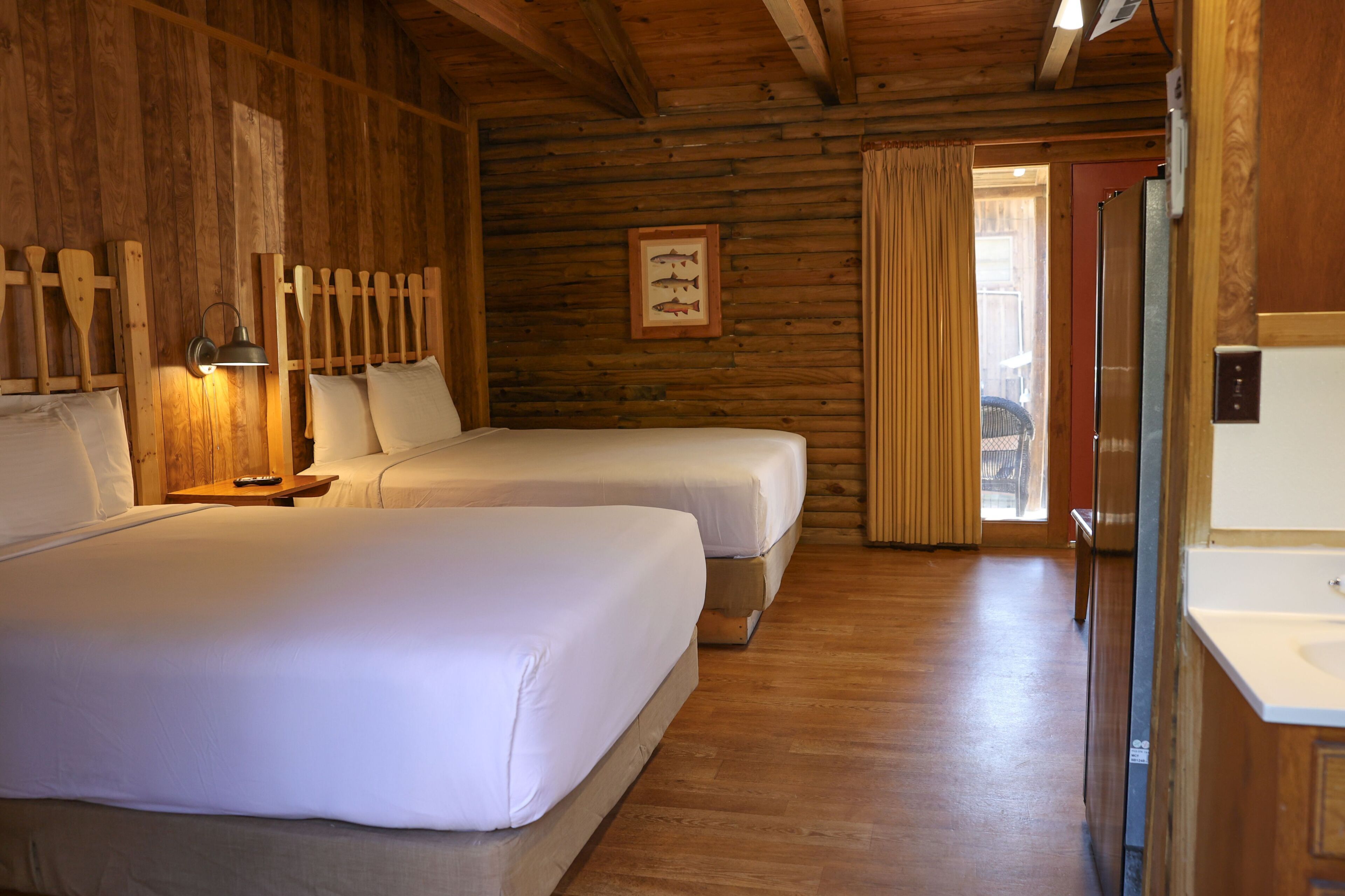 Ein rustikal eingerichtetes Zimmer mit zwei Betten, einer Holzbalkendecke, einem Bild von einem Fisch im Rahmen sowie einem Badezimmer mit Waschbecken und Spiegel.