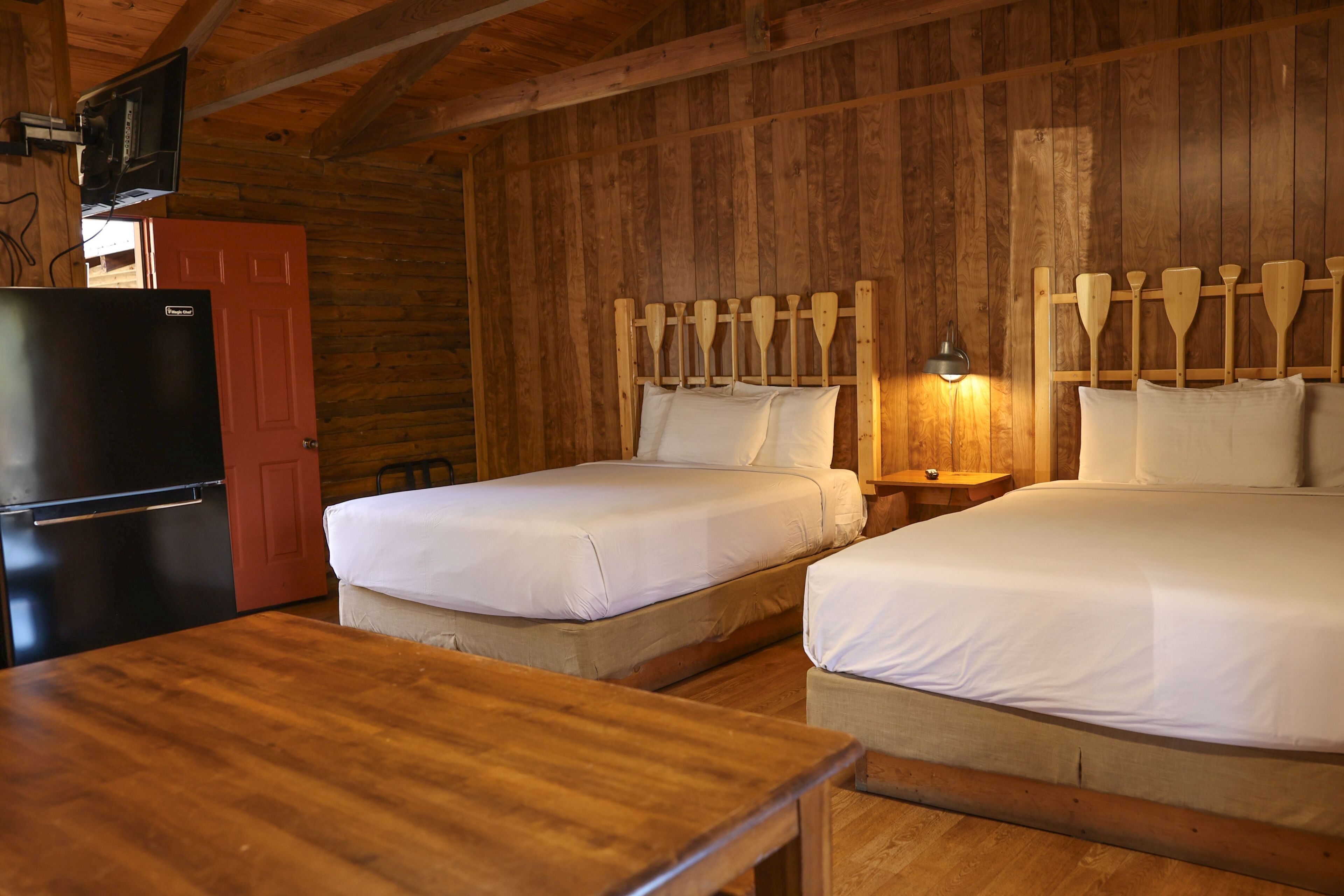 A cabin-style room with two beds, a wooden table, a red door, and a black refrigerator.
