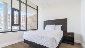 A bedroom with a large window, a bed, a nightstand, and a view of buildings outside. - Unit 3 - Modern Fully Furnished 3 Bedroom Apartment (Toronto)