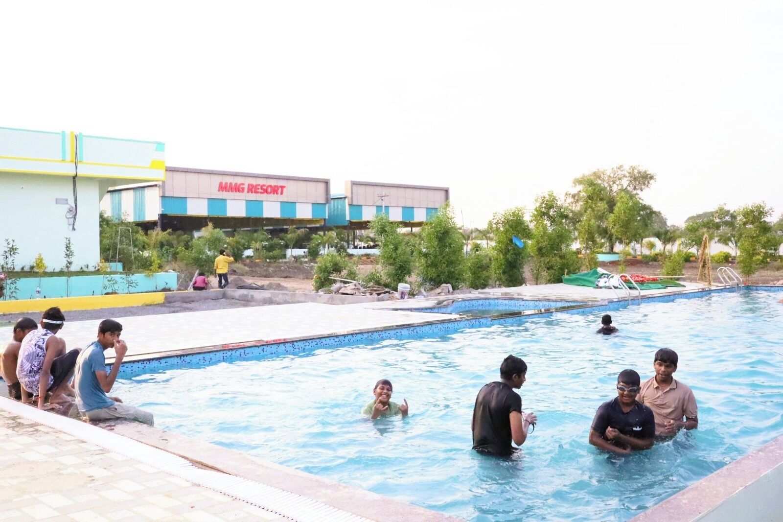 A swimming pool area with people in the water and on the edge, a building labeled 'MMG RESORT' in the background, and some greenery around.