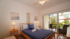 Une chambre à coucher avec un lit, des lampes de chevet, une tête de lit en bois, une fenêtre donnant sur un balcon et un ventilateur de plafond.