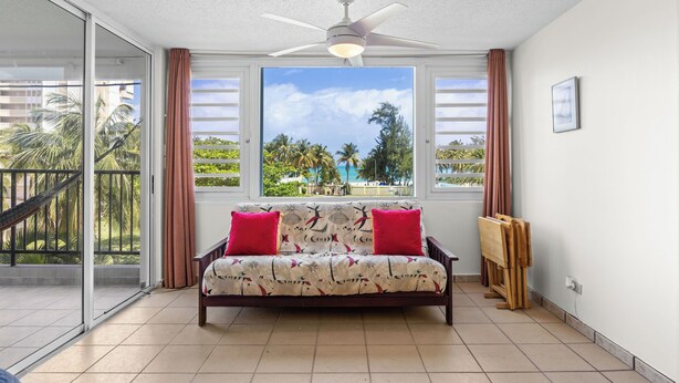 A room with a sofa, a ceiling fan, a large window with a view of palm trees, and a balcony with a hammock.
