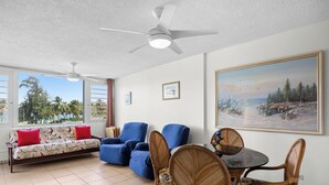 A living room with a sofa, two blue armchairs, a dining table with chairs, a ceiling fan, and a large landscape painting on the wall.