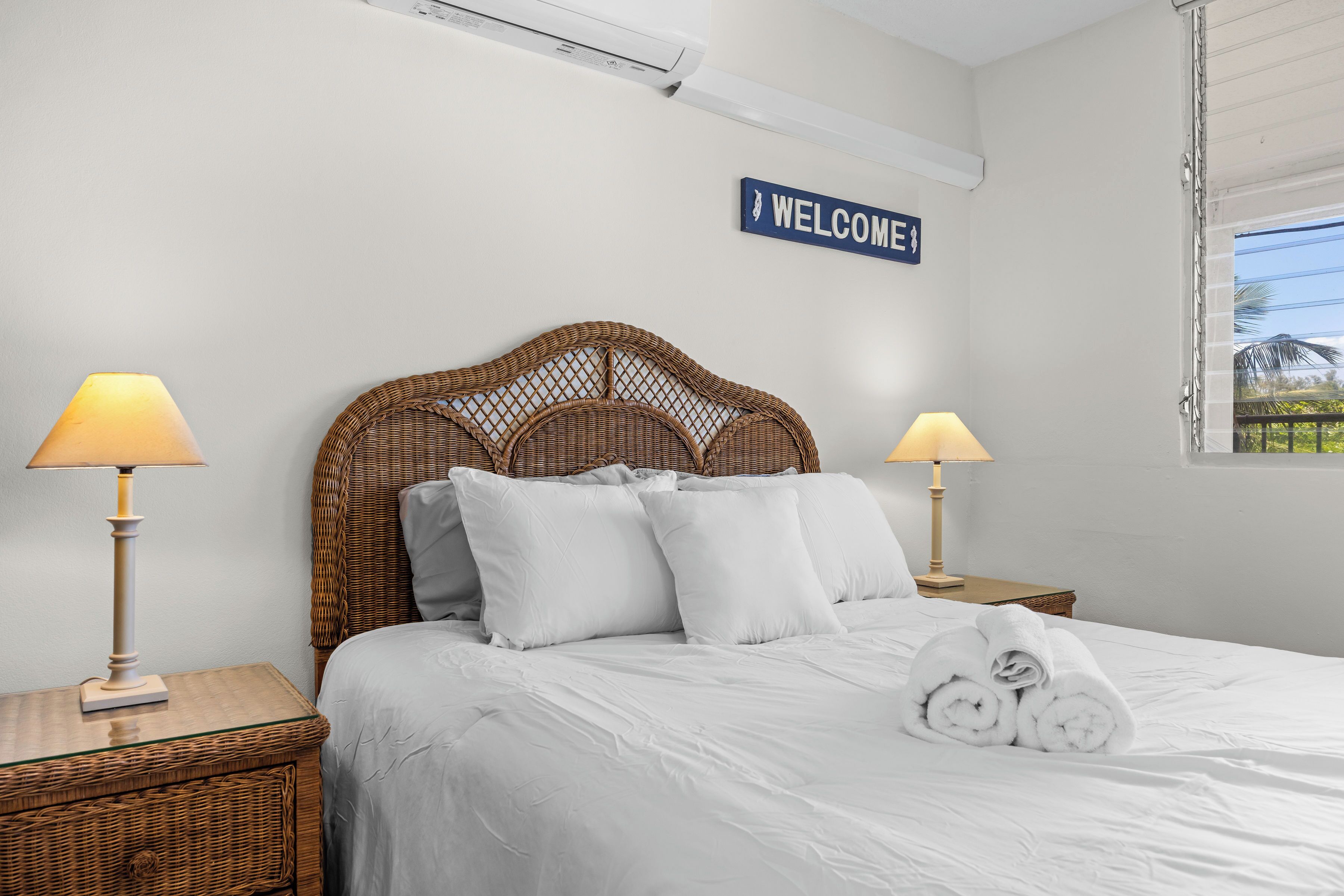 A neatly made bed with white linens, two bedside lamps, a wicker headboard, and a 'WELCOME' sign on the wall.