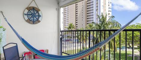Un balcon avec un hamac, une décoration murale en forme de boussole, une table en verre avec une bougie, et une vue sur de hauts immeubles et des palmiers.