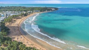 An aerial view of a coastal area with a beach, clear turquoise waters, and a dense forested area.