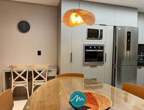 A modern kitchen with a microwave, oven, and refrigerator, a wooden dining table with chairs, and a wall clock. - Urban Gameleira - Miramar Hospedagens (João Pessoa)