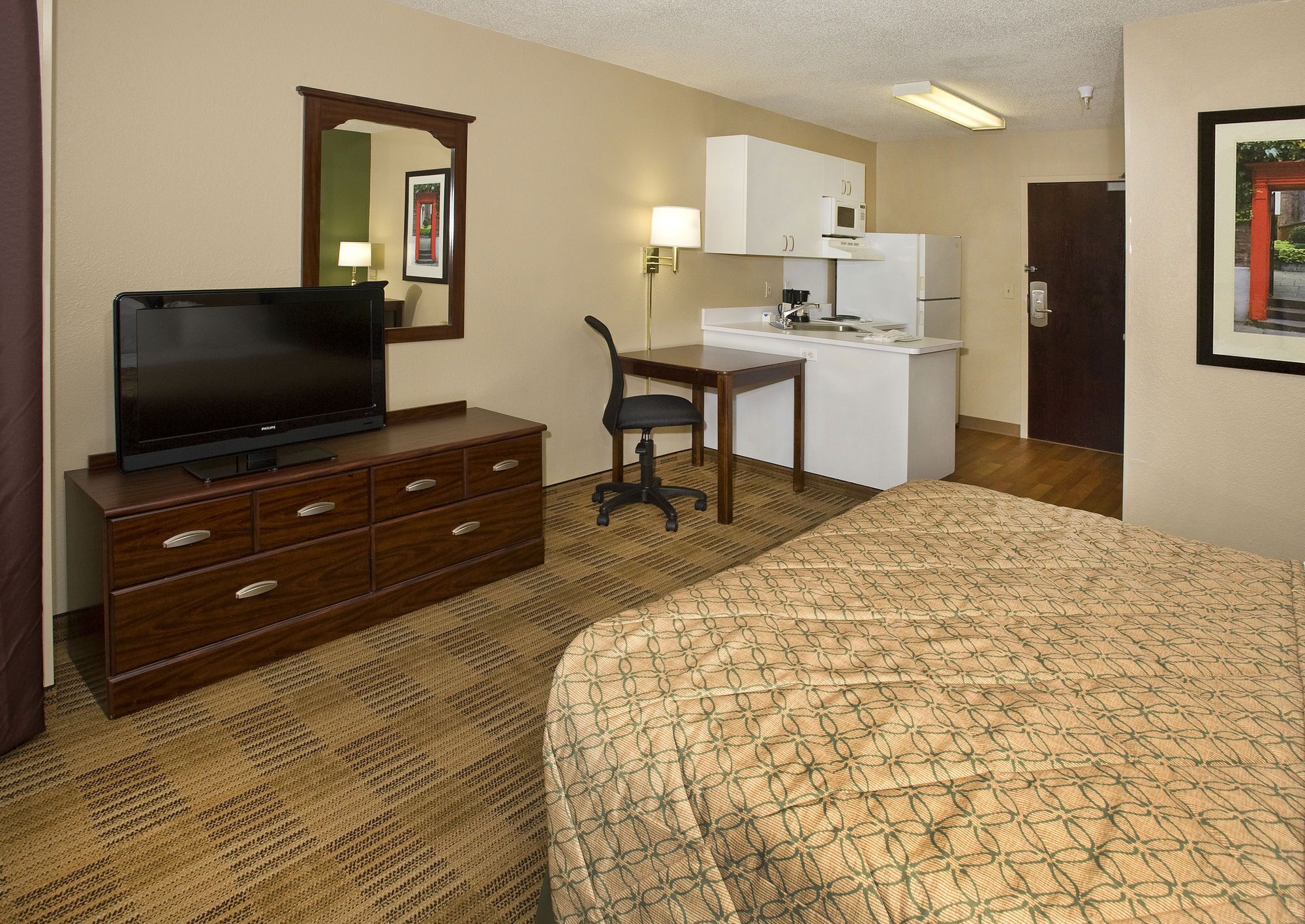 A hotel room with a bed, a TV on a dresser, a desk with a chair, a kitchenette, and a door.