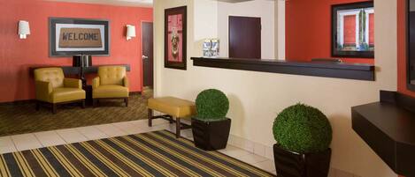 A hotel lobby with a reception desk, two yellow armchairs, a striped carpet, and two potted plants.