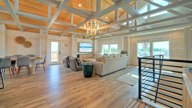 A spacious living area with a wooden floor, a sofa, a dining table with chairs, and a staircase with a railing. - Third Sandbar & Seaglass Landing by Avantstay. Sleeps 30 2 Homes + Dual Pools + Outdoor Living Steps to Beach (Port Aransas)