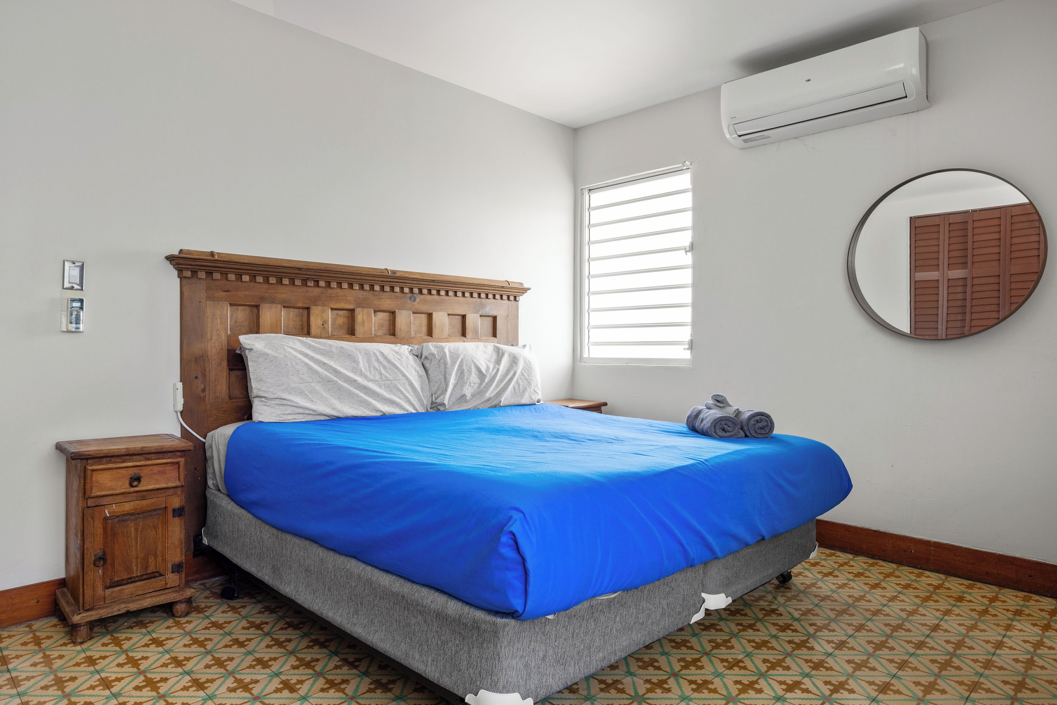 A bedroom with a wooden headboard, a blue bedspread, a bedside table, a wall-mounted air conditioner, a window with blinds, and a round mirror.