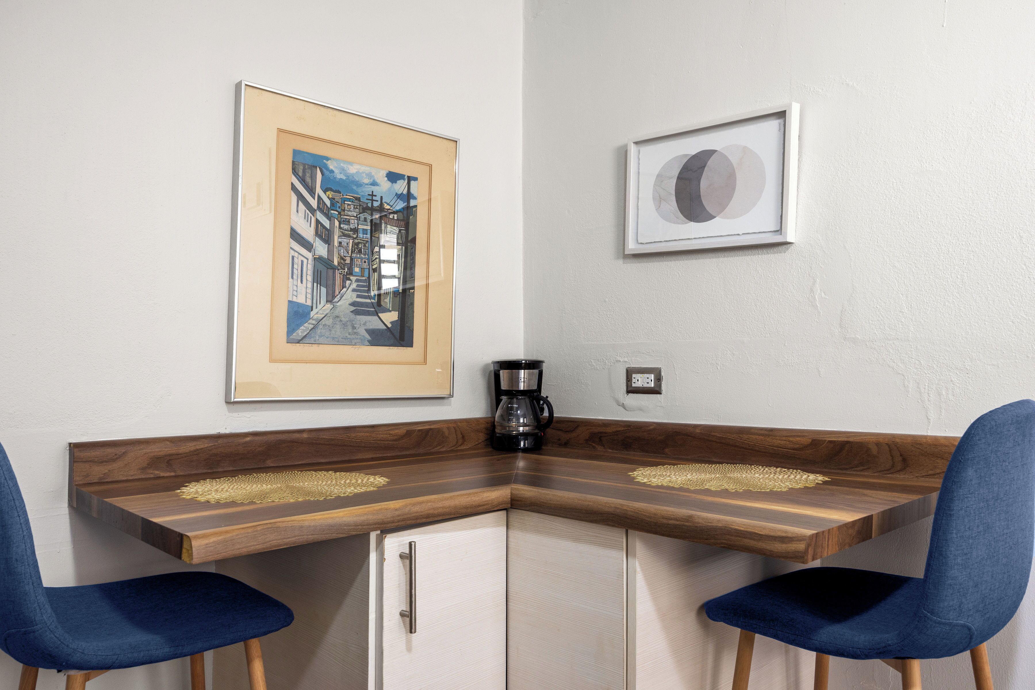 Ein Schreibtisch an der Ecke mit zwei Stühlen, einer Kaffeemaschine und zwei gerahmten Bildern an der Wand.