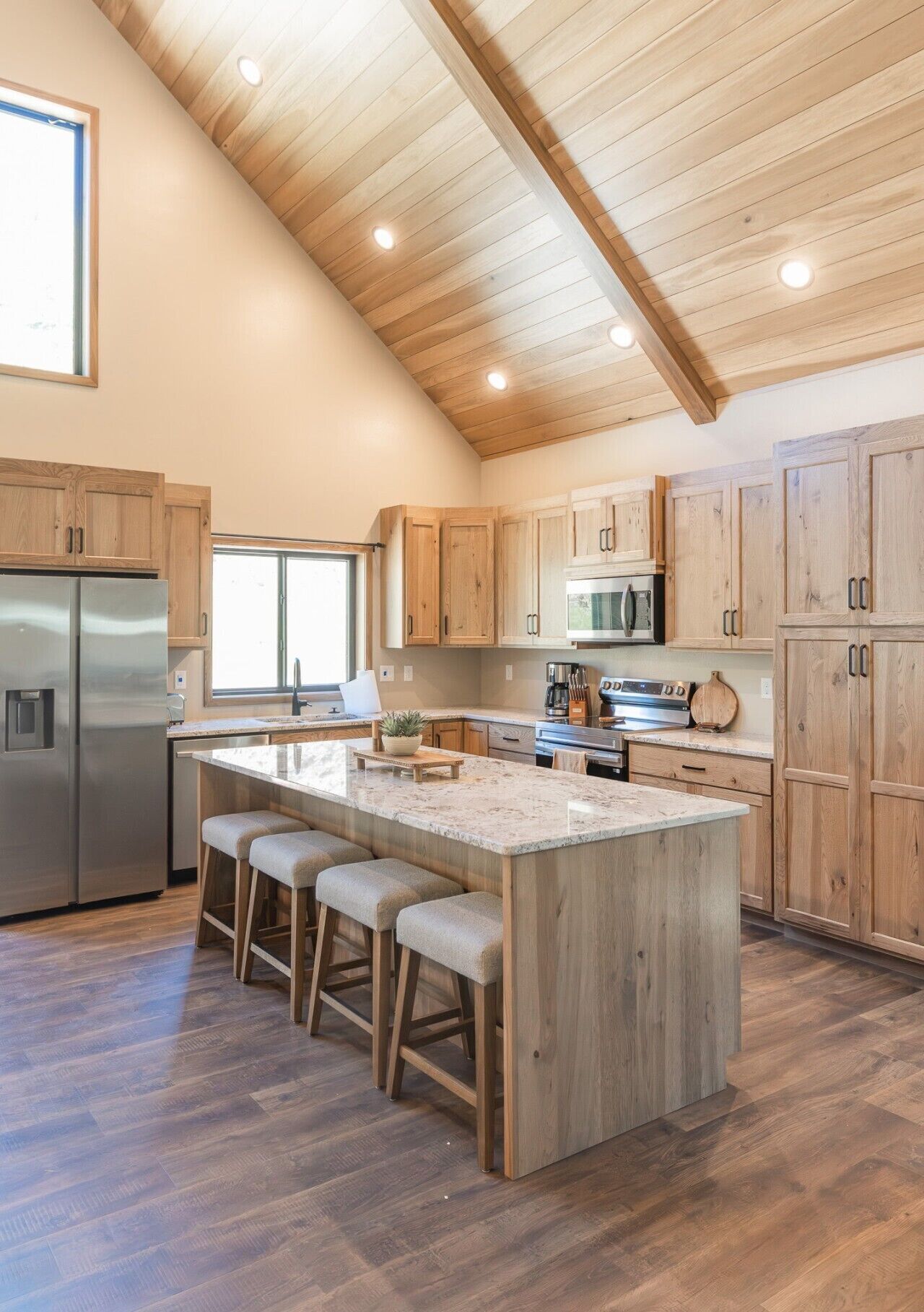A modern kitchen with wooden cabinets, a stainless steel refrigerator, a large island with bar stools, and a wooden ceiling.