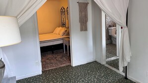 Un dormitorio con cama, una lámpara de noche, un colgador de macramé en la pared y un baño visible a través de una puerta abierta.