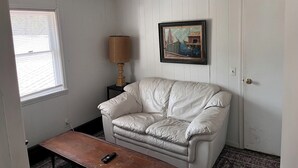 Una habitación con un sillón de cuero blanco, una mesita, una lámpara y un cuadro en la pared.