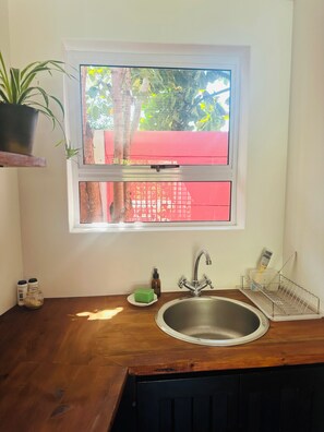 A kitchen with a window, a sink, a plant, and a red fence outside. - Flamingo Palm House - boutique-inspired stay (Cape Town)