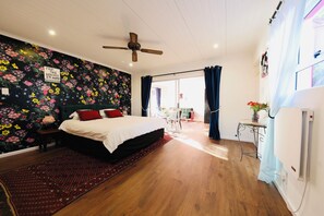 A bedroom with a bed, a ceiling fan, a window with curtains, a small table with a plant, and a wall with a floral mural. - Flamingo Palm House - boutique-inspired stay (Cape Town)