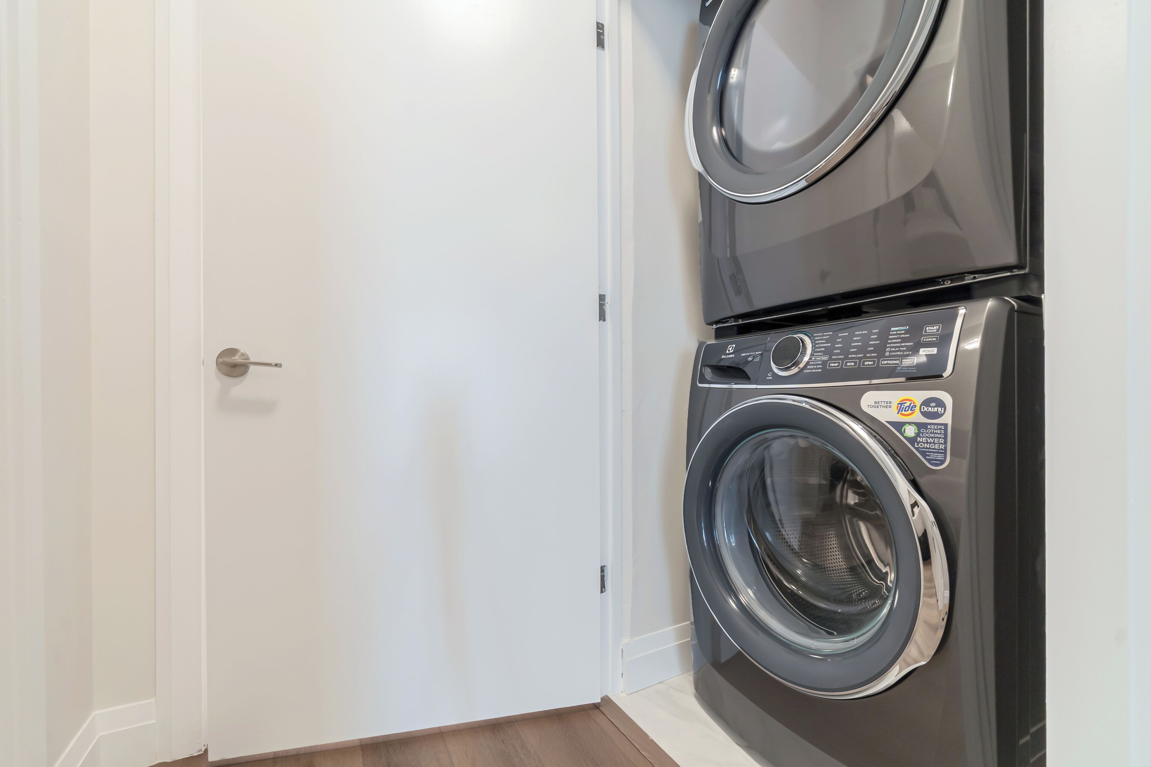 Une buanderie équipée d’un lave-linge et d’un sèche-linge superposés, d’une porte blanche et d’un plancher en bois.