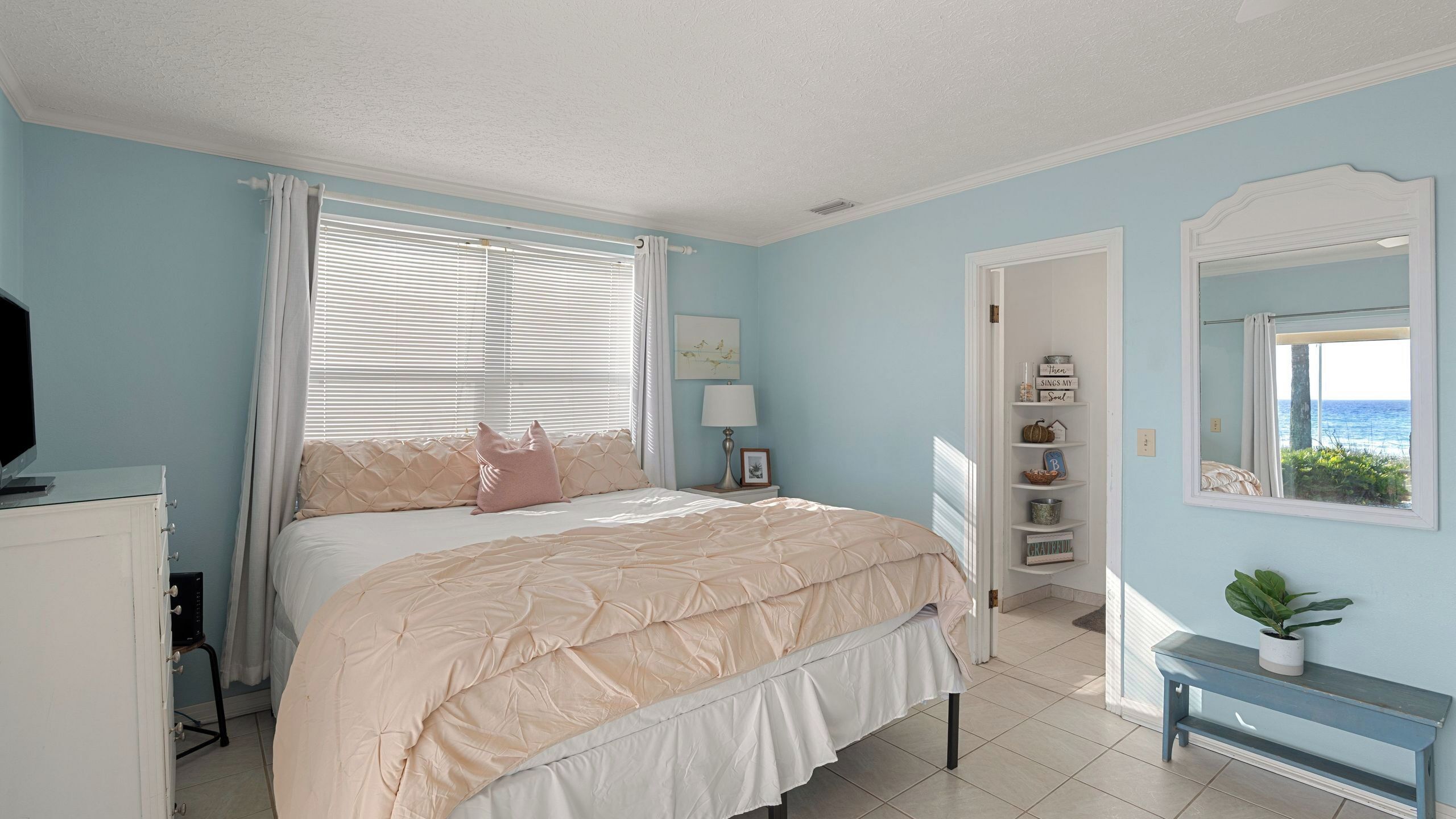A bedroom with a bed, a bedside table with a lamp, a TV, a window with blinds, a door leading to another room, and a view of the ocean.