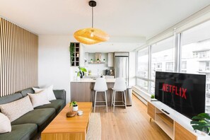 A modern living room with a sofa, coffee table, and TV. There is a kitchen area in the background with a view of the city. - Olympic Village Oasis |Downtown Van |Free Parking (Vancouver)
